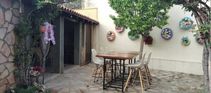 Una terrazza esterna con pavimento in pietra, un tavolo di legno e sedie. Sono presenti piante e fiori in vaso sulla parete.