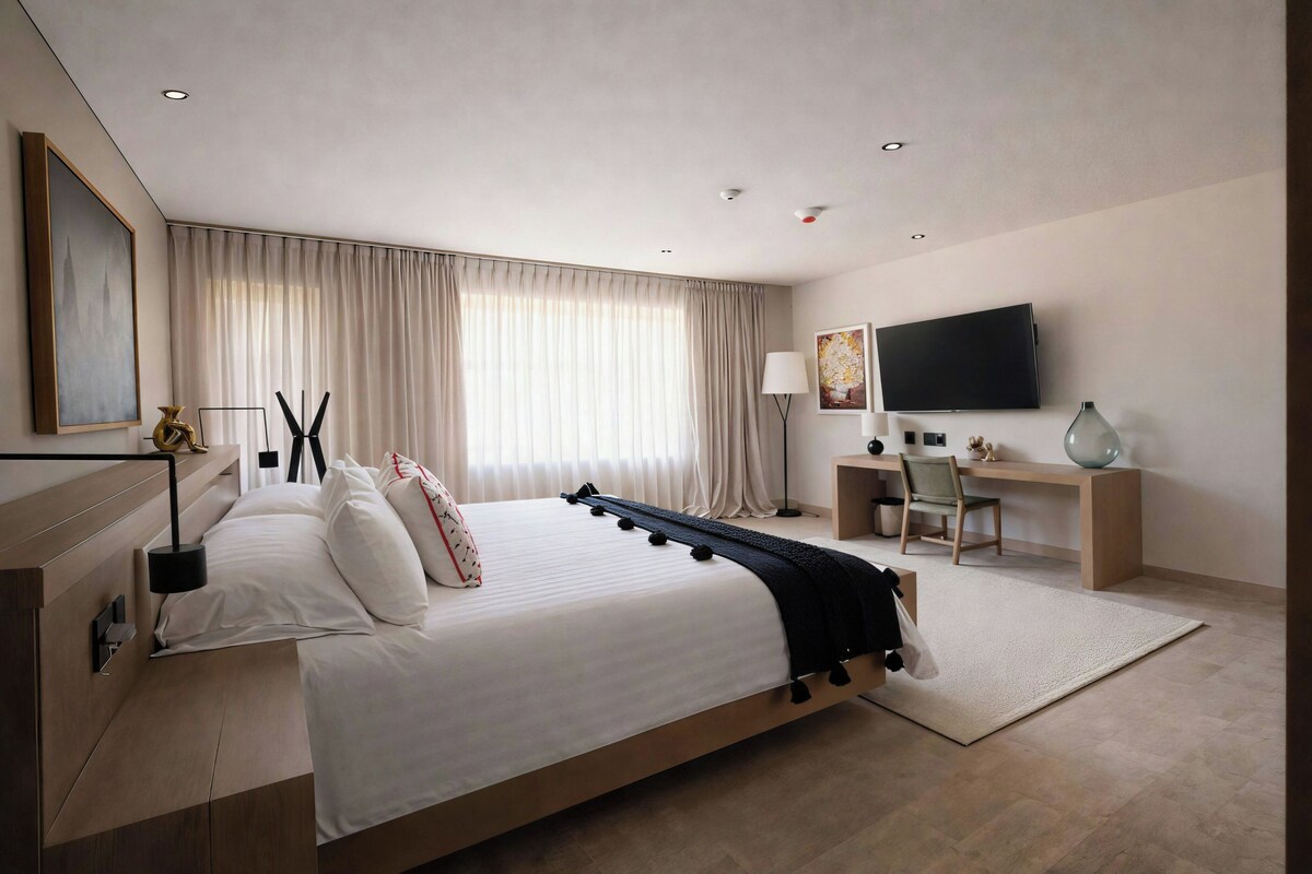 Quarto de hotel moderno com uma cama grande, uma escrivaninha com cadeira, televisão de tela plana e uma janela ampla com cortinas.