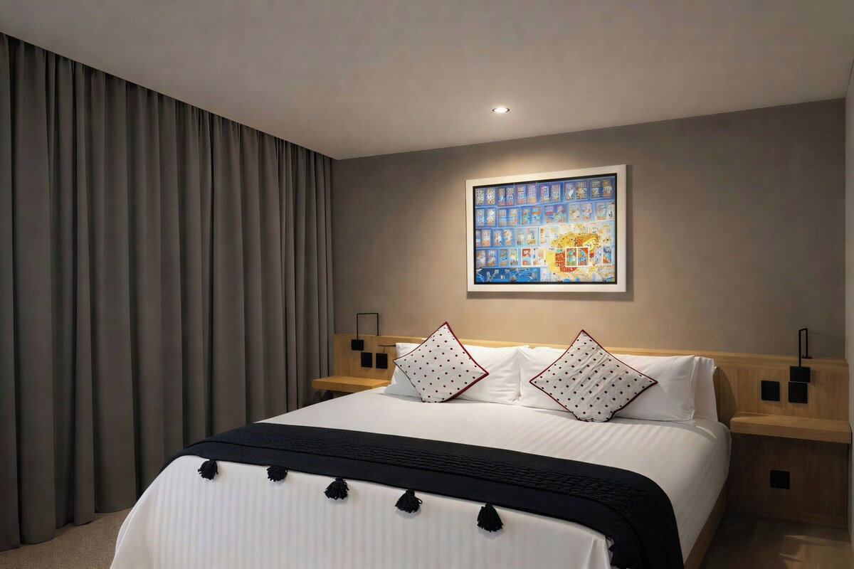 Quarto de hotel com uma cama grande, cabeceira com prateleira embutida, duas mesas de cabeceira, uma obra de arte na parede e uma janela ampla com cortinas.