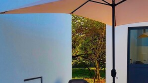 Un espacio al aire libre con dos sillas, una mesa, un paraguas grande y una puerta a un edificio.
