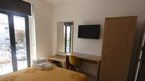 Habitación de hotel con cama, escritorio, silla, armario, televisión y ventana con vista a coches estacionados y un edificio.