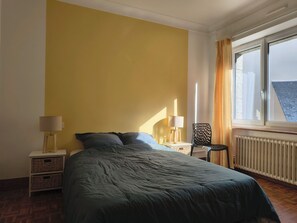 Ein Schlafzimmer mit Bett, Nachttisch, Stuhl, Fenster mit Vorhängen und Heizkörper.