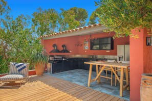 Een buitenkeuken met een eettafel en stoelen, omringd door een houten terras en plantenpotten.