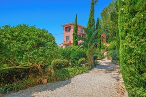 A gravel pathway leading to a building with a pink facade, surrounded by lush greenery and tall trees. - Villa Arcadia: Sleeps 14, 5x11 Pool, Ocean View, Near the Beach, Petanque, Ping-Pong (Carqueiranne)