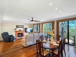 A spacious living room with a fireplace, a television, a ceiling fan, and a dining area with a table set for four. - Villa Magenta - 2-Storey Villa at Cypress Lakes Resort (POKOLBIN)