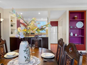 A dining area with a table set for two, a vase with flowers, and a kitchen in the background. - Villa Magenta - 2-Storey Villa at Cypress Lakes Resort (POKOLBIN)