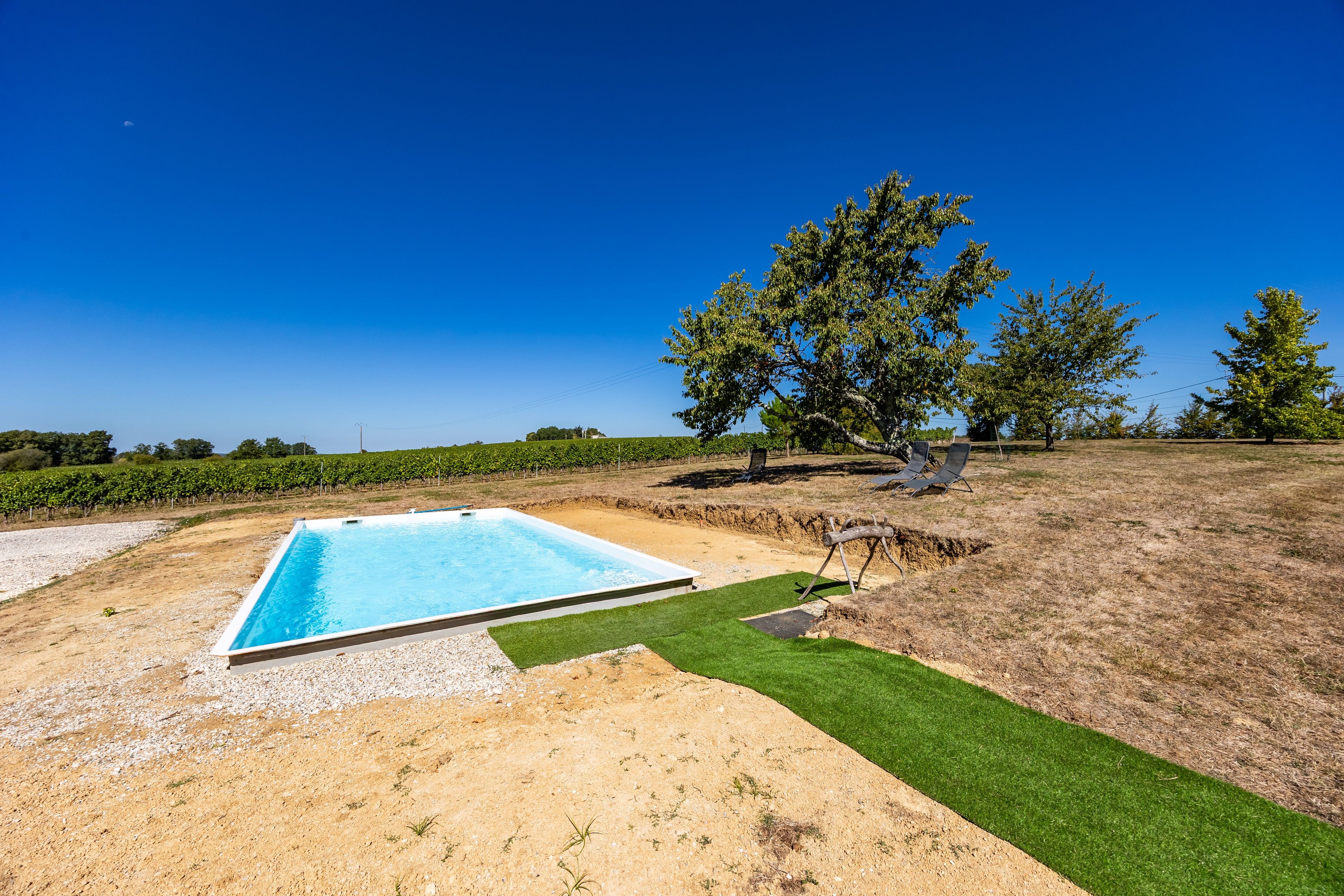 A rectangular swimming pool with a clear blue water surface, surrounded by a sandy area and a patch of green artificial turf.