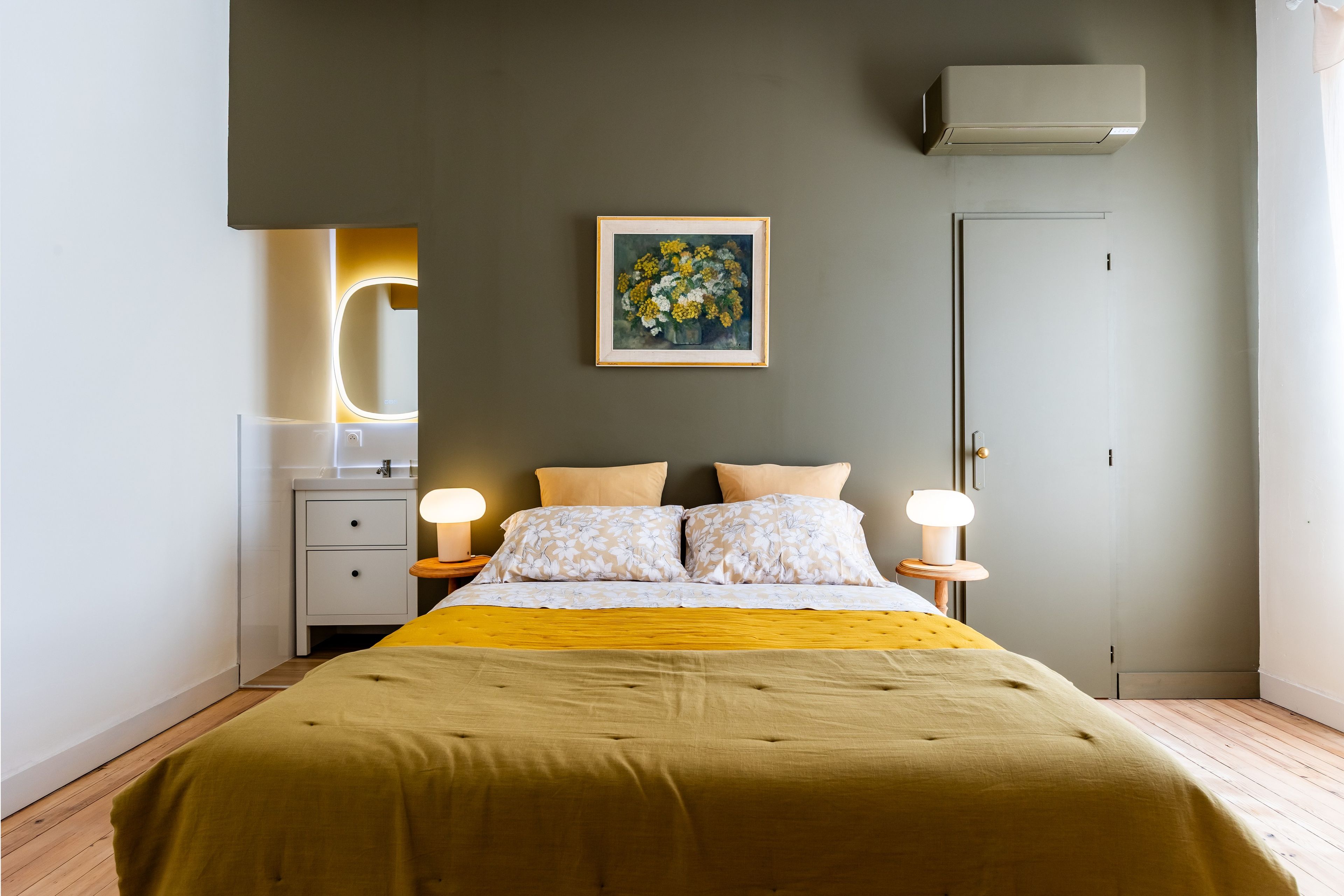 A bedroom with a bed, bedside lamps, a nightstand, a wall-mounted air conditioner, and a framed painting.