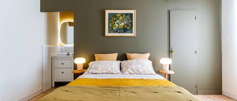 A bedroom with a bed, bedside lamps, a nightstand, a wall-mounted air conditioner, and a framed painting.