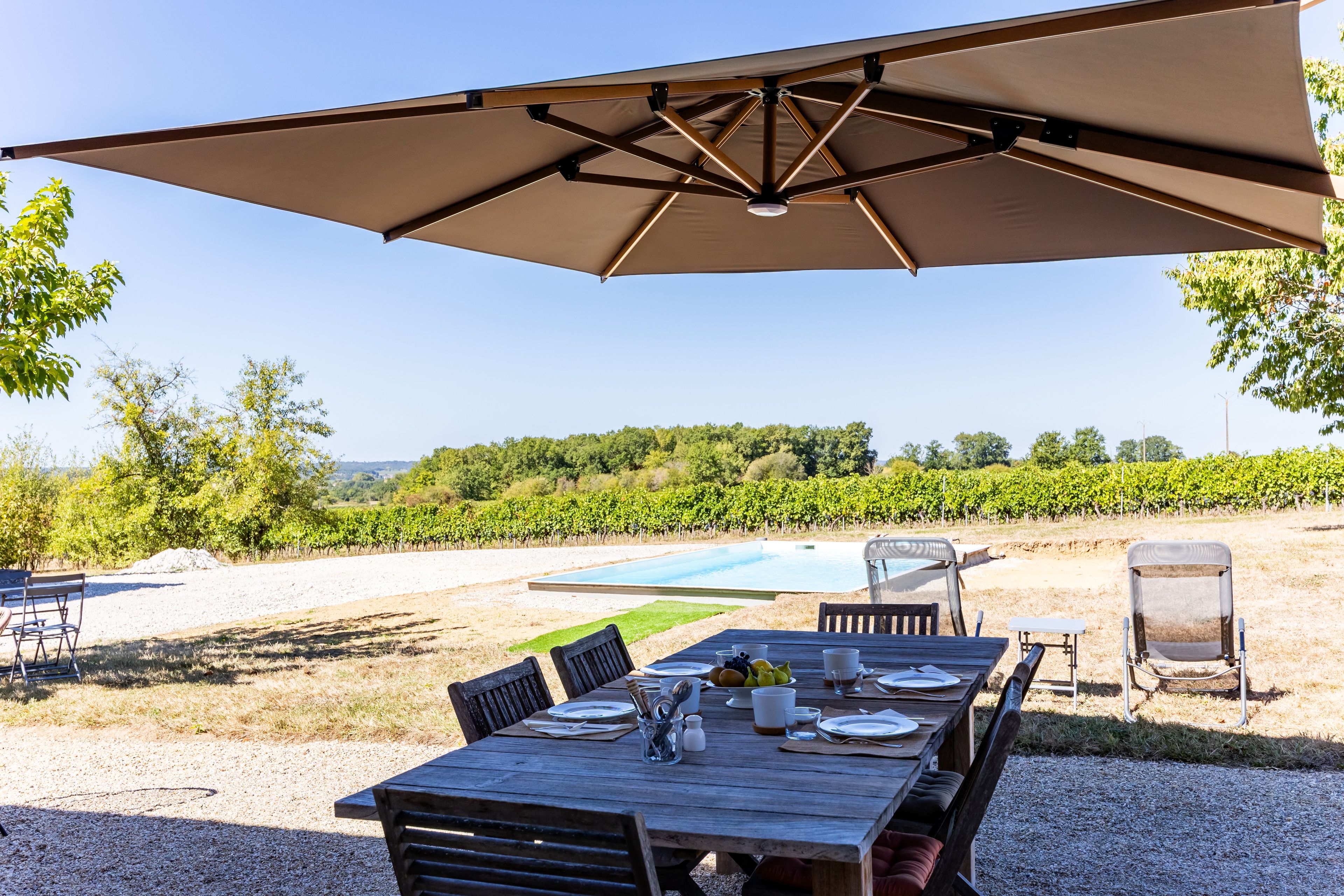 Un espace extérieur aménagé pour les repas, avec une table dressée pour quatre personnes, un grand parasol et, en arrière-plan, une vue sur un vignoble et une piscine.
