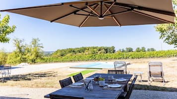An outdoor dining area with a table set for four, a large umbrella, and a view of a vineyard and pool in the background.