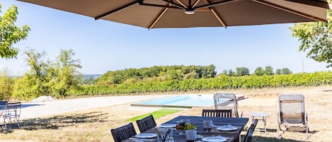 Ein Essbereich im Freien mit einem Tisch für vier Personen, einem großen Sonnenschirm und einem Blick auf einen Weinberg und Pool im Hintergrund.