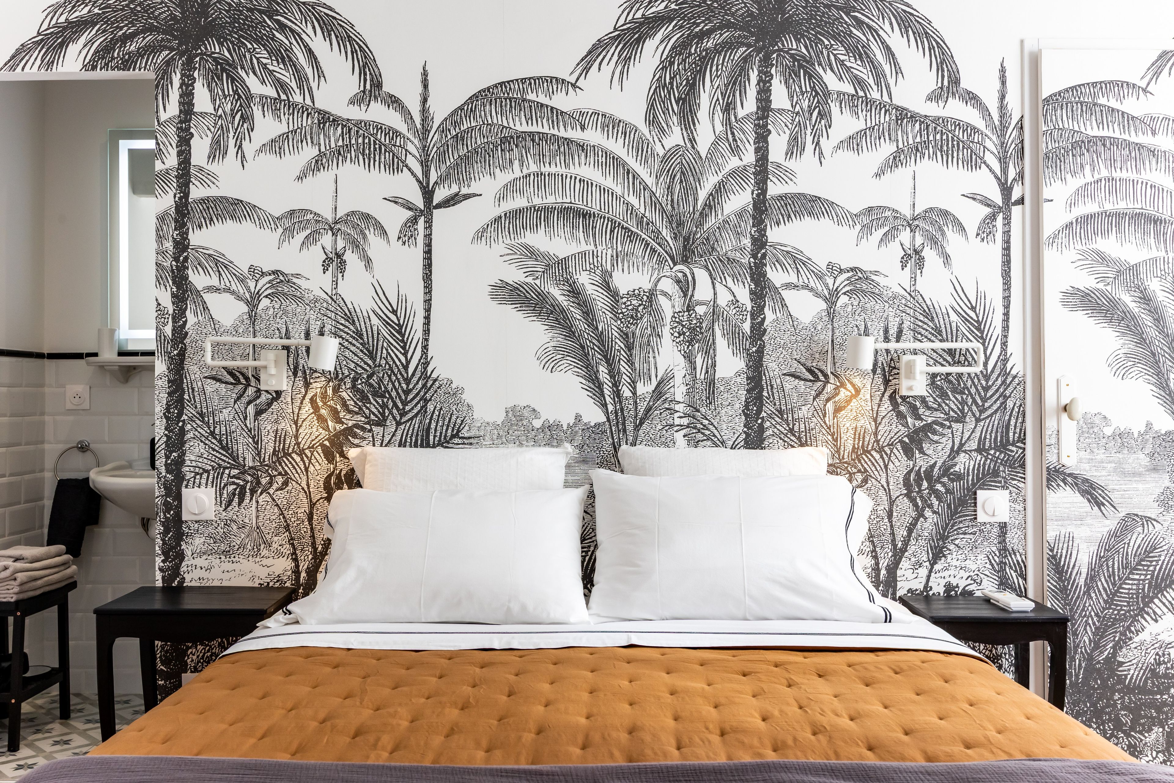 A bedroom with a bed, white pillows, a tufted headboard, a nightstand, a lamp, and a wall with a tropical plant mural.