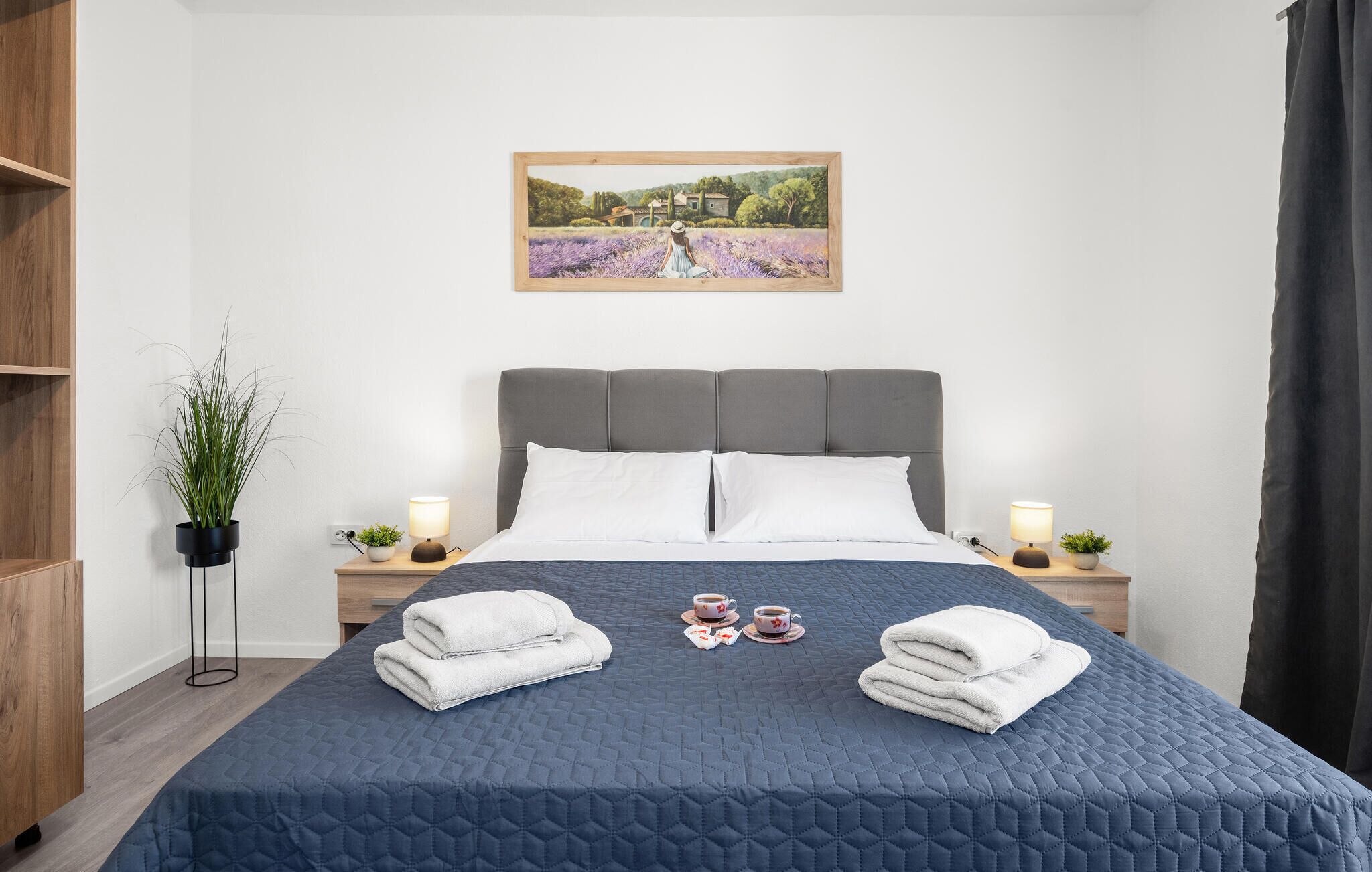 Ein ordentlich bezogenes Bett mit einer blauen Stegdecke, zwei Nachttische mit Lampen, ein gerahmtes Bild an der Wand und ein Blumentopf links.