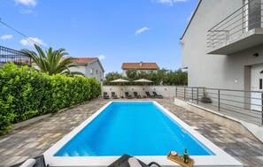 Ein Pool mit Liegestühlen und Sonnenschirmen, umgeben von Grünflächen und Gebäuden.