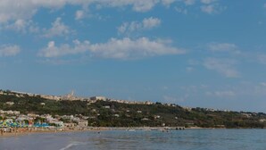 Una playa costera con cielo despejado, colinas al fondo y algunas embarcaciones en el agua.