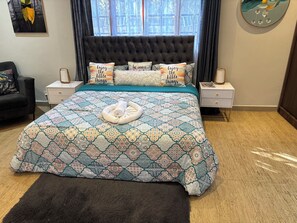 Una cama bien tendida con ropa de cama y almohadas estampadas, una mesita de noche con una lámpara, y vistas al exterior a través de una ventana.