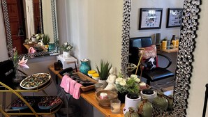 Ein Friseursalon mit einem großen Spiegel, Regalen mit diversen Gegenständen und einem Tisch mit Telefon und Tasche.