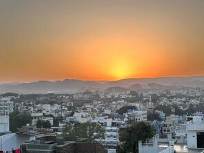 A cityscape at sunset with buildings and distant mountains. - Stayostel (Udaipur)