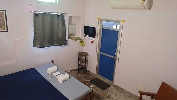 Uma cama de solteiro com colchão azul, uma pequena mesa com uma cadeira, um ventilador de teto, uma televisão e uma porta com moldura azul.