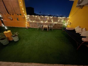 A rooftop terrace with artificial grass, a seating area with chairs and a table, a decorative fence with lights, and a view of the cityscape at night. - Stayostel (Udaipur)