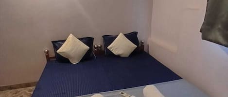 A single bed with a blue mattress, two pillows, and a wooden headboard. There are towels on the bed and a small table with a lamp.