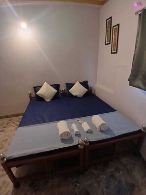 A single bed with a blue mattress, two pillows, and a wooden headboard. There are towels on the bed and a small table with a lamp. - Stayostel (Udaipur)