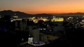 Paisagem urbana ao anoitecer, com edifícios iluminados e montanhas ao longe.