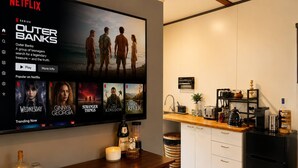 A modern living room with a large flat-screen TV displaying a Netflix interface, a wooden coffee table with decorative items, and a kitchen area with a sink and countertop appliances.