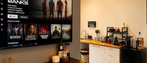 Una sala de estar moderna con un televisor de pantalla plana grande que muestra la interfaz de Netflix, una mesa de centro de madera con objetos decorativos, y una zona de cocina con un lavabo y electrodomésticos en la encimera.