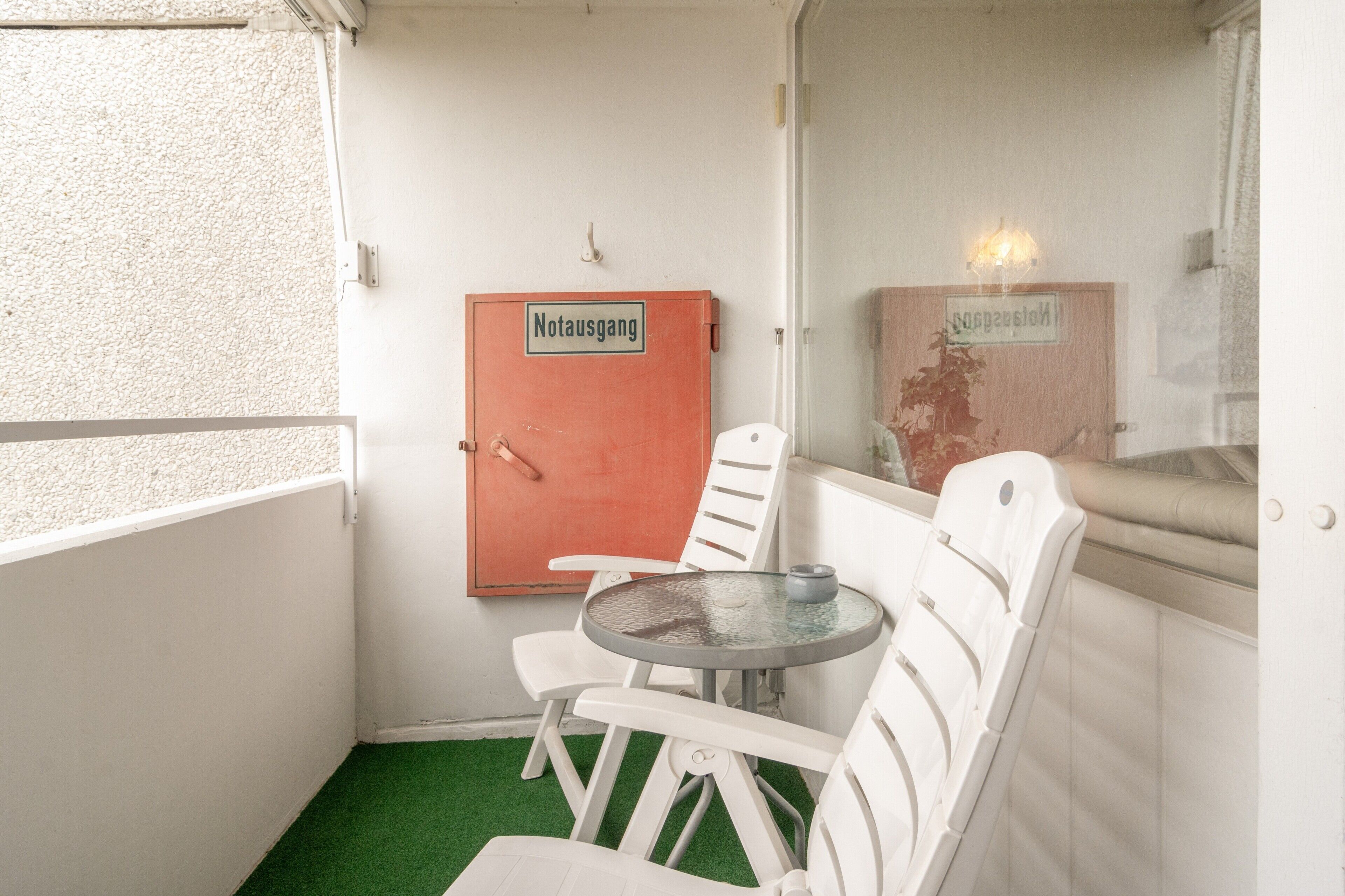 Una stanzaetta con una porta bianca con la scritta 'Notausgang', una sedia bianca, un piccolo tavolo rotondo e un pavimento in erba sintetica verde.