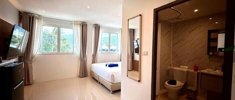 A hotel room with a bed, a television, a bathroom with a toilet and sink, and a view of greenery outside.