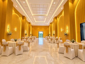 Une salle de banquet où les tables sont disposées en rangées, chacune étant agrémentée de chaises blanches et de rubans dorés, et une grande porte se trouve au fond de la salle.