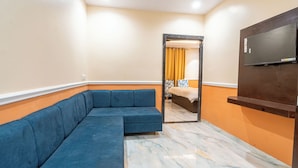 A room with a sectional sofa, a bed with a yellow curtain, a television mounted on the wall, and a marble floor. - Shree Maa Vaishno Hotel & Resort (Ujjain)