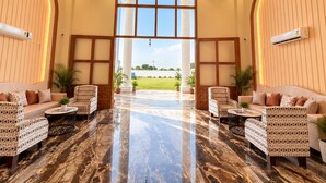 A spacious lobby with a marble floor, two sofas with patterned upholstery, a large window with a view of a green field, and an air conditioning unit mounted on the wall.