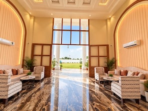A spacious lobby with a marble floor, two sofas with patterned upholstery, a large window with a view of a green field, and an air conditioning unit mounted on the wall. - Shree Maa Vaishno Hotel & Resort (Ujjain)