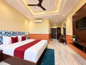 A hotel room with a bed, a ceiling fan, an air conditioning unit, a TV, a desk, and a mirror. - Shree Maa Vaishno Hotel & Resort (Ujjain)