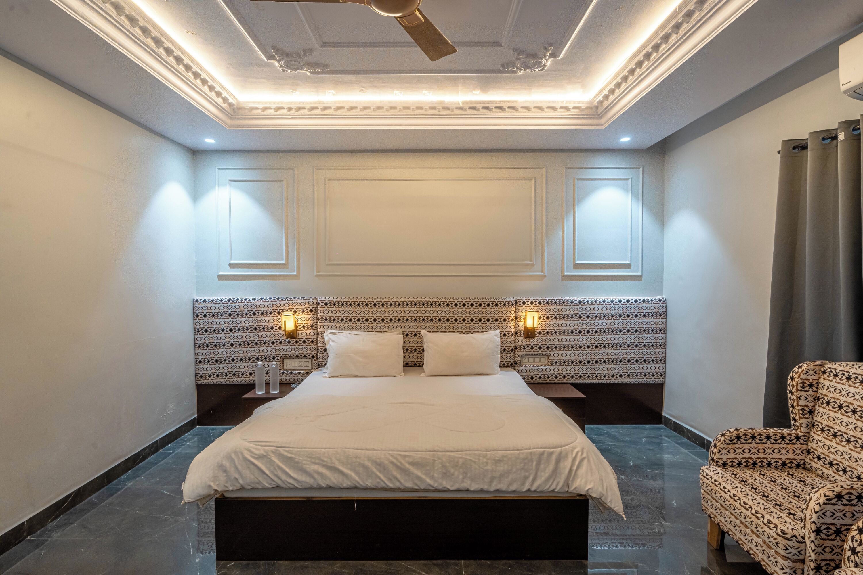 A hotel room with a large bed, a patterned headboard, two bedside lamps, and a patterned armchair.