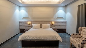 A hotel room with a large bed, a patterned headboard, two bedside lamps, and a patterned armchair. - Shree Maa Vaishno Hotel & Resort (Ujjain)
