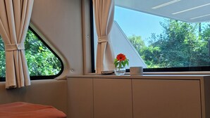 Una habitación con cama, ventana con cortinas, un jarrón con una flor y vista a la vegetación exterior.