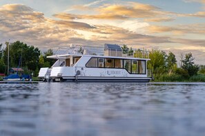 Ein weißes Boot mit der Aufschrift 'LUXBOOT', liegt an einer grünen Uferböschung, im Hintergrund sind Bäume zu sehen.