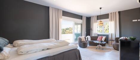 A modern bedroom with a large bed, a seating area with a table, and a view of the outdoors.