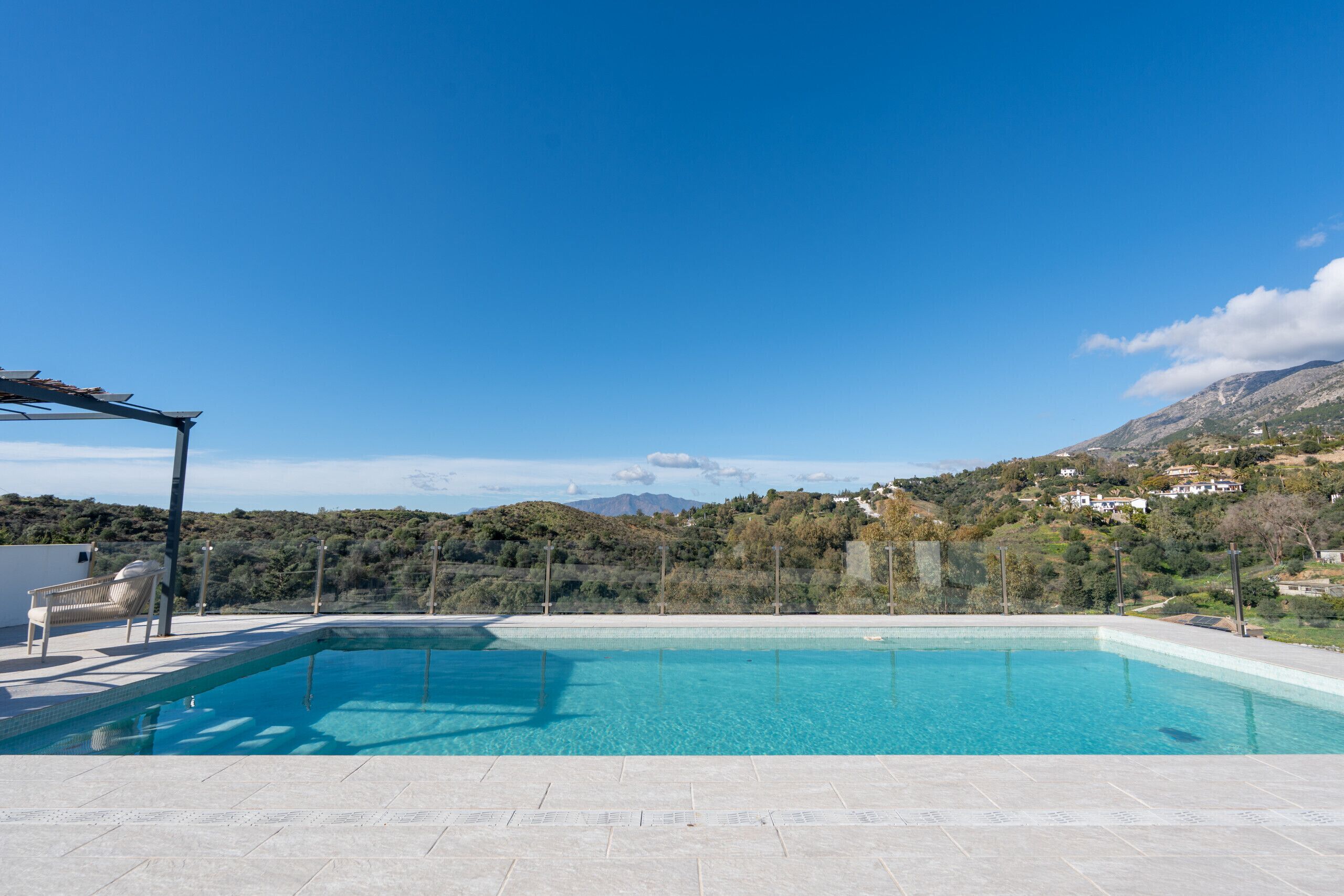 Ein klares, blaues Schwimmbecken mit einer Pergola und einem Liegestuhl, umgeben von einer gebirgigen Landschaft.