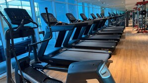 En række løbebånd i et fitnesscenter med store vinduer og trægulv.