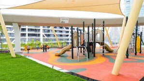 Un patio de recreo al aire libre con diversas estructuras para jugar, un área techada y una vista de un edificio moderno al fondo.
