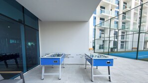 Et udendørs område med to fodboldborde, siddepladser og en tennisbane, der kan ses gennem en glasvæg.