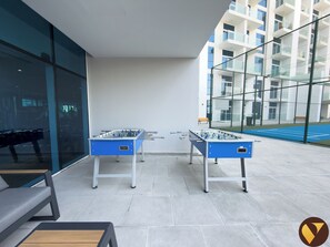 An outdoor area with two foosball tables, seating, and a tennis court visible through a glass wall. - Vogue | Skyz by Danube near Dubai Miracle Garden (Dubai)