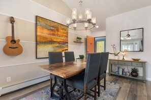 A dining room with a wooden table, four chairs, a chandelier, a guitar, and a large abstract painting. - 1 Car Garage | Gourmet Kitchen | On Bus Route (Steamboat Springs)