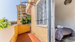 A narrow balcony with a tiled floor, a bed with a grey duvet, and a window with a metal grid. - THAT'S AMORE FIUMICINO AIRPORT  (Fiumicino)