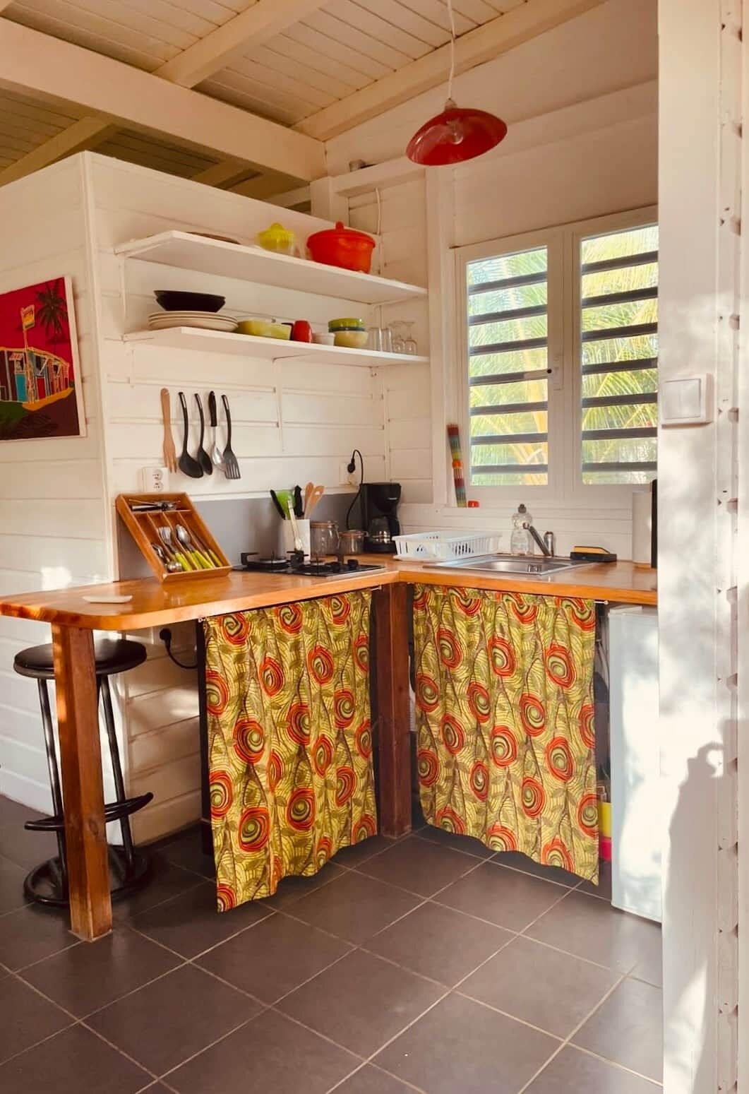 Uma cozinha com bancada de madeira, dois bancos, uma luminária pendente vermelha e uma janela com persianas.